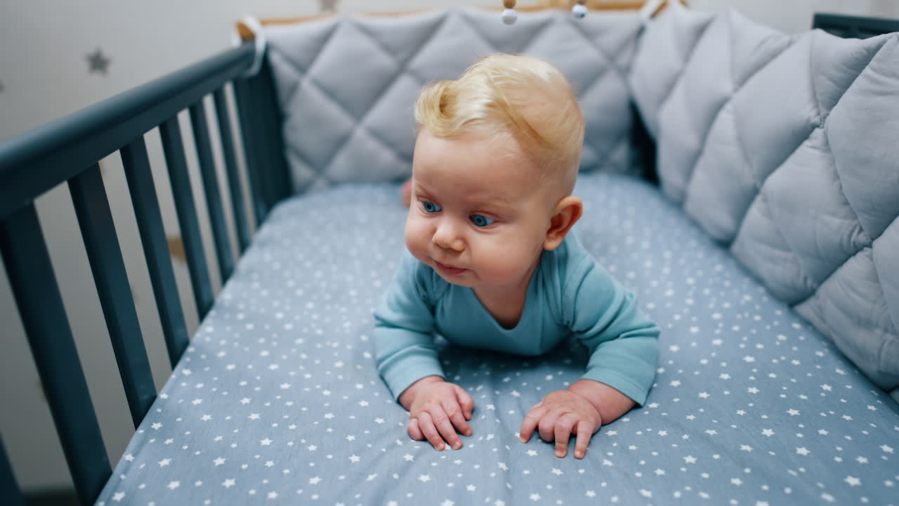 Cute baby boy lies in his crib. Blue-eyed blond child looking around. Close up.