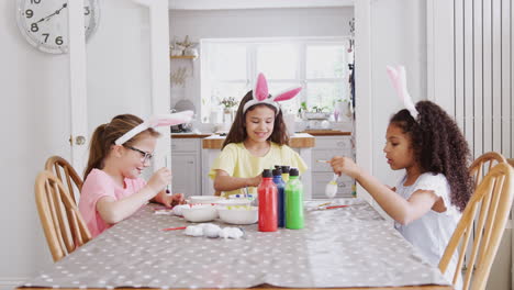 group of girls wearing bunny ears sitting at table decorating eggs for easter at home