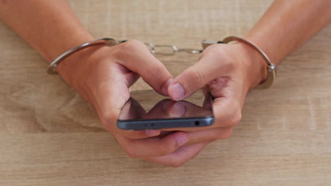 Hands in handcuffs using smartphone on wooden table for Cyber security Concept