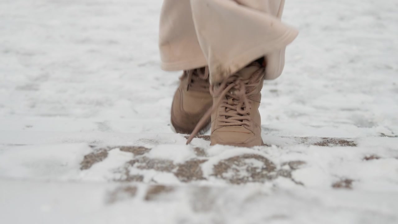 Vista en ángulo bajo de la parte inferior de las piernas con pantalones anchos beige y botas marrones con cordones, caminando por una acera nevada y compacta, mostrando un paso hacia adelante, un paso elevado, contacto del talón, aguanieve, tracción invernal y frío.