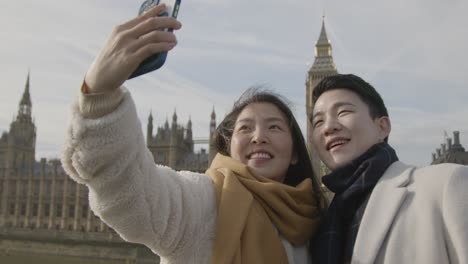 young asian couple on holiday posing for selfie in front of houses of parliament in london uk