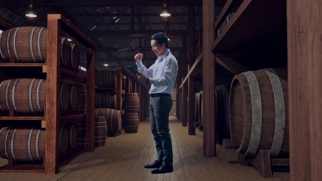 full body side view of an asian male professional worker standing with his tablet in cellar of winery, he raises his fist up with screaming goal