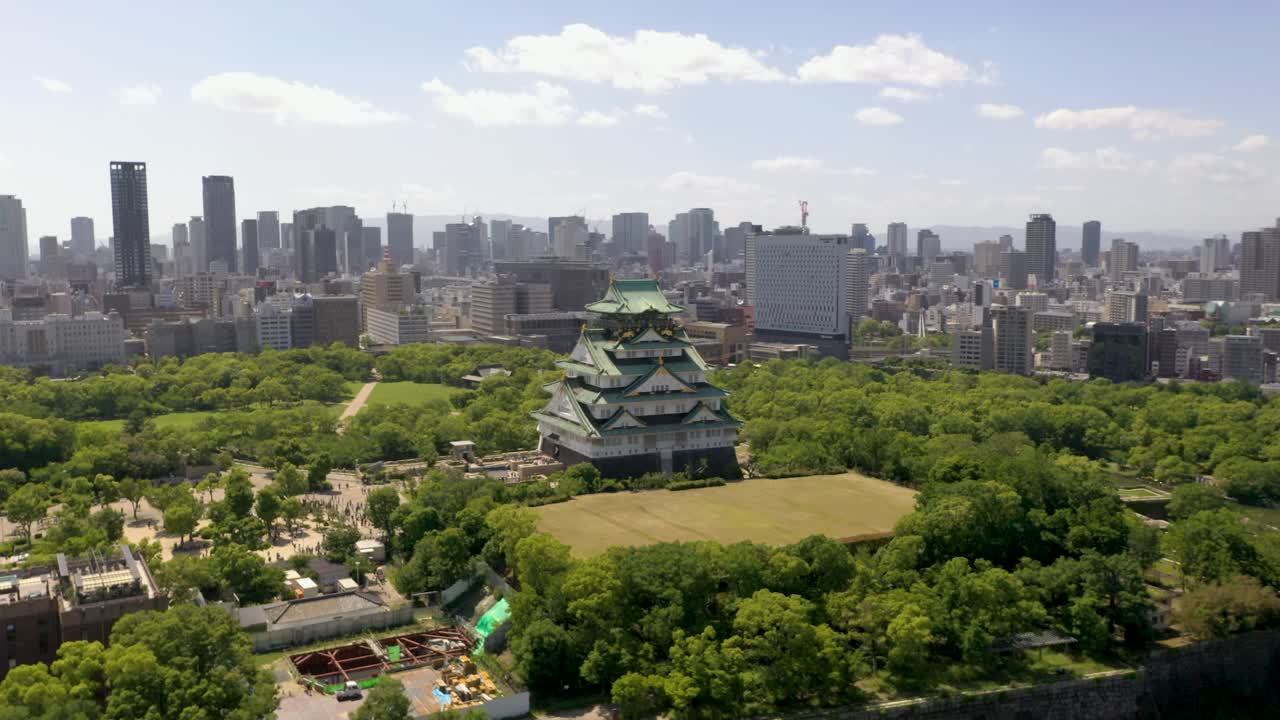 Aerial spinning around historic Osaka Castle with park, moat, skyscraper, and city in Osaka, Japan