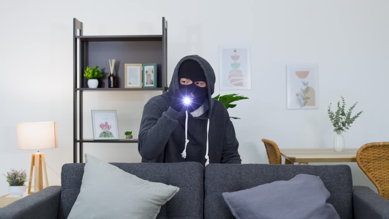A masked intruder in a hoodie uses a flashlight to search a dark living room