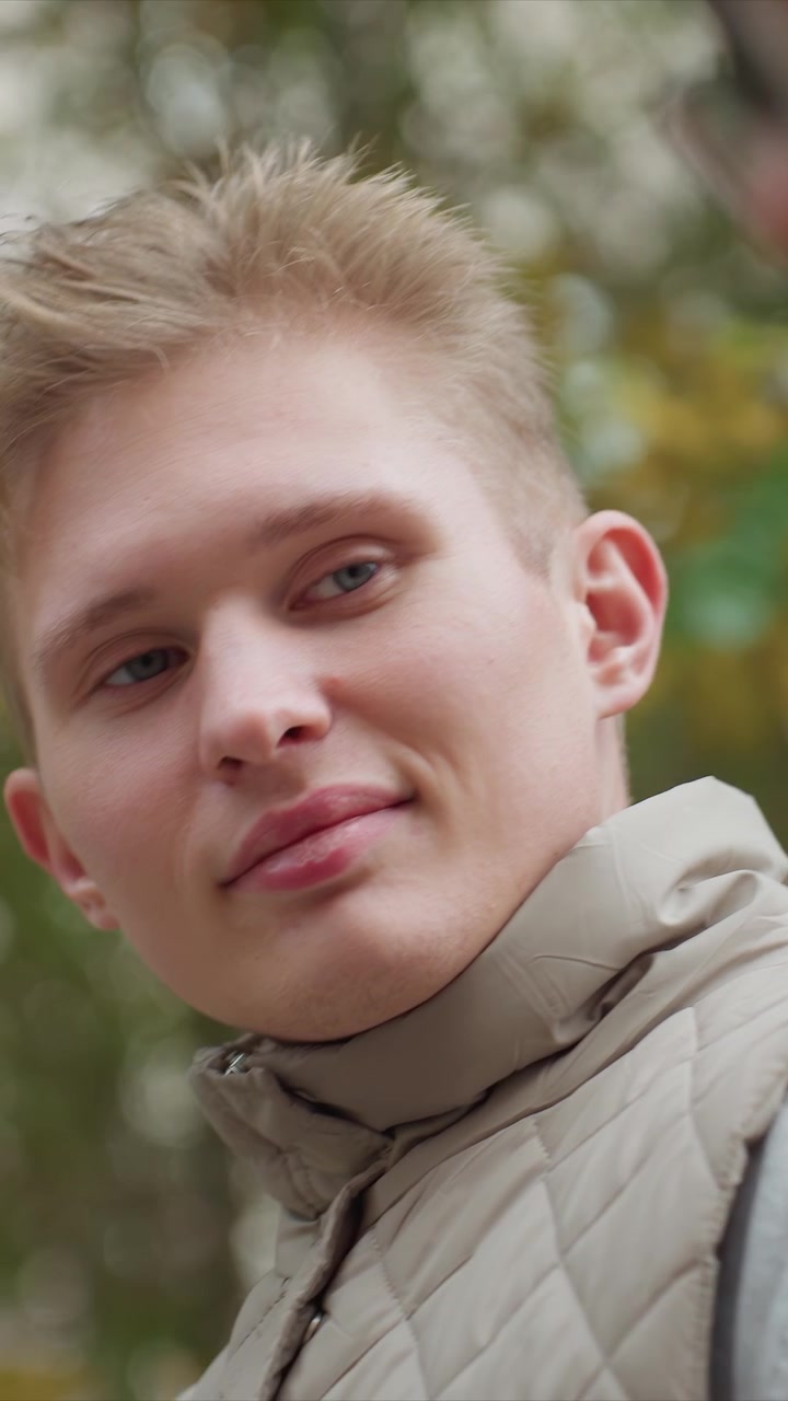 Profil mladého muže v béžové prošívané vestě, který mluví. Osoba se pohybuje minimálně, s lehkými posuny hlavy. Jednotné pozadí s rozostřenými stromy naznačuje
