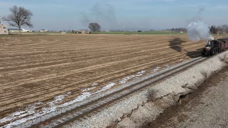 a steam train chugs along empty tracks lined with fields, showcasing picturesque farmland and blue skies. wisps of smoke billow from the steam engine as it moves forward