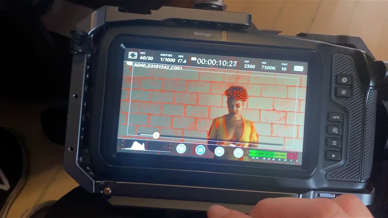 A videographer starts playback on her cinema camera of a woman in a yellow dress with a candle against a white brick wall