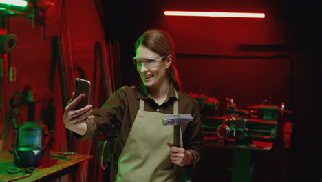 Caucasian-welder-woman-in-goggles-and-holding-hammer-making-a-video-call-on-smartphone-in-metal-workshop