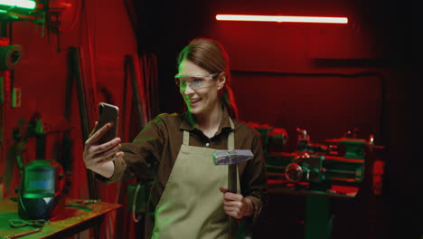 caucasian welder woman in goggles and holding hammer making a video call on smartphone in metal workshop