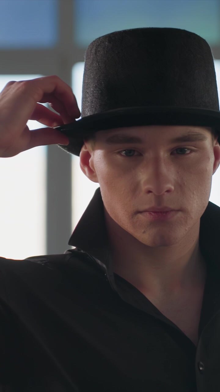 Primo piano di un uomo con camicia scura e cappello nero che si sistema. Tatuaggio visibile sul collo, una luce naturale soffusa crea un'atmosfera delicata. Ambiente interno con sfondo sfocato che suggerisce un interno.