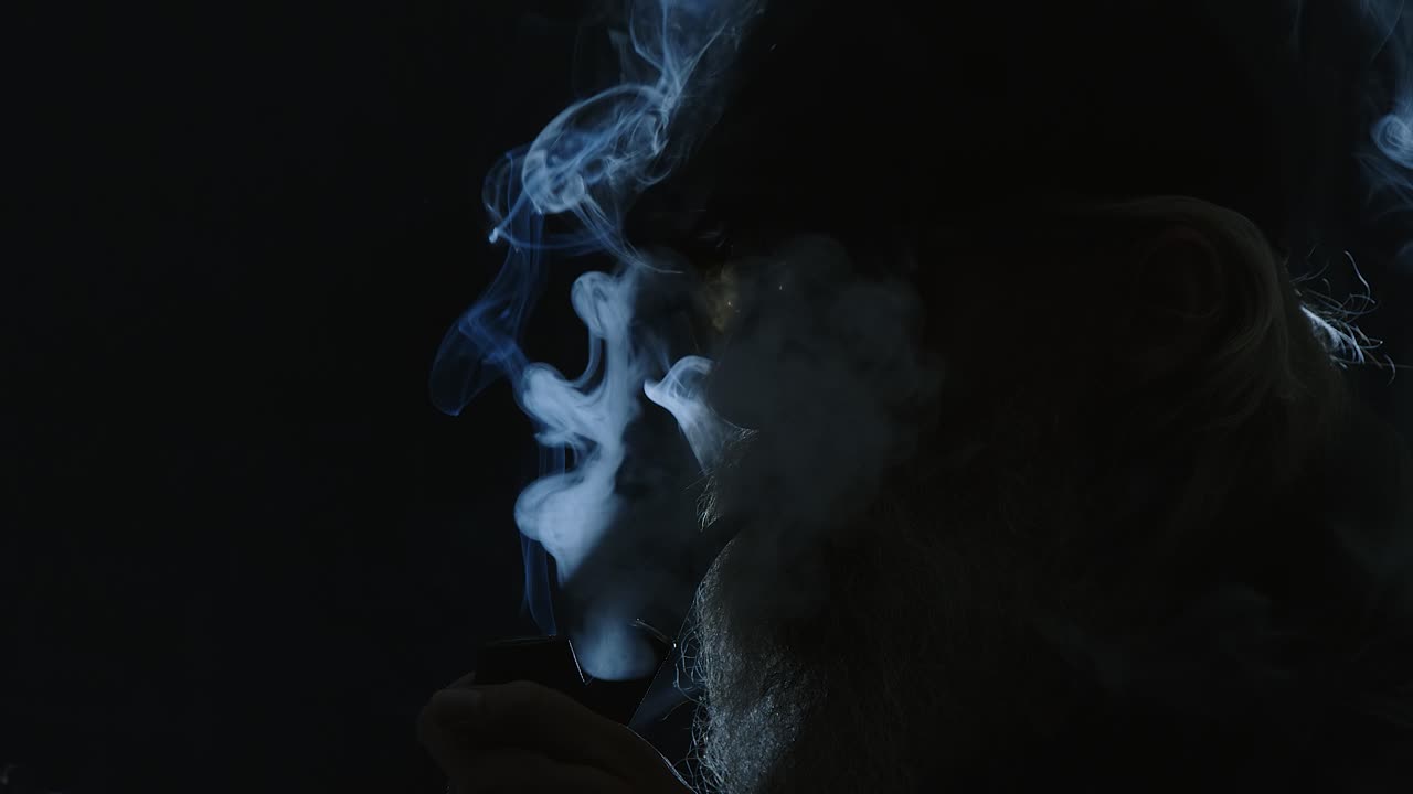 Backlit bearded male in flat cap lights pipe in black room, close-up