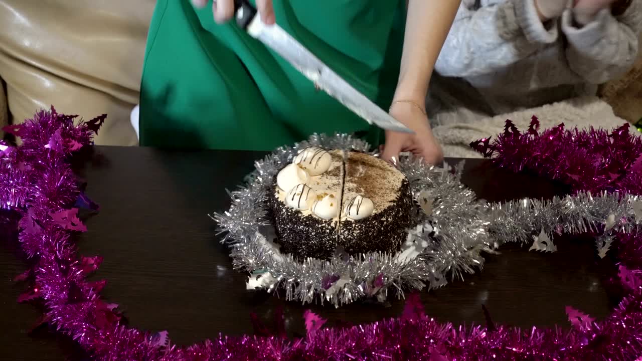 Children clap their hands and cut the chocolate cake into pieces with a large knife on the table decorated with tinsel during the holiday home. Close up. The view from the top. 4K.