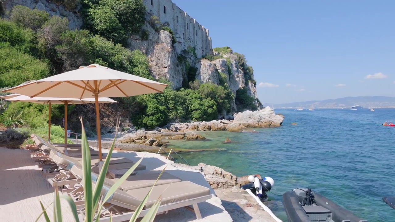 Lettini e ombrelloni da beach club sotto le scogliere di Fort Royal sopra acque turchesi, Ile Sainte-Marguerite, Francia