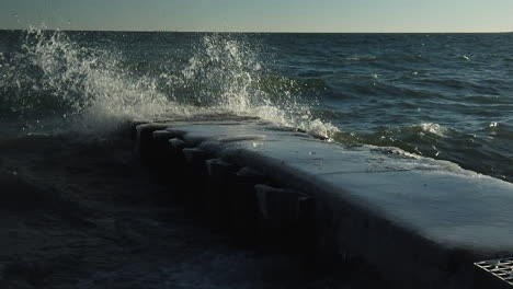 Wide-shot-of-winter-waves-colliding-and-splashing-in-slow-motion-onto-an-ice-covered-jetty
