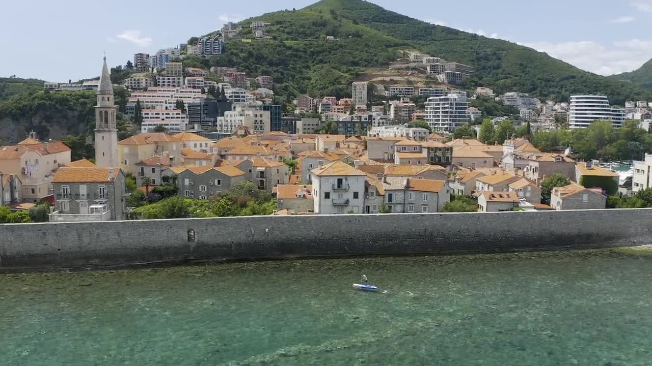 Vista aerea con drone della Città Vecchia di Cattaro con mura in pietra lungo il lungomare Adriatico, edifici con tetti arancioni, un'alta torre campanaria e la città collinare in Montenegro.