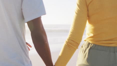 Smiling-african-american-couple-walking-and-holding-hands-on-sunny-beach