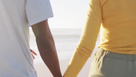 una pareja afroamericana sonriente caminando y tomándose de las manos en una playa soleada