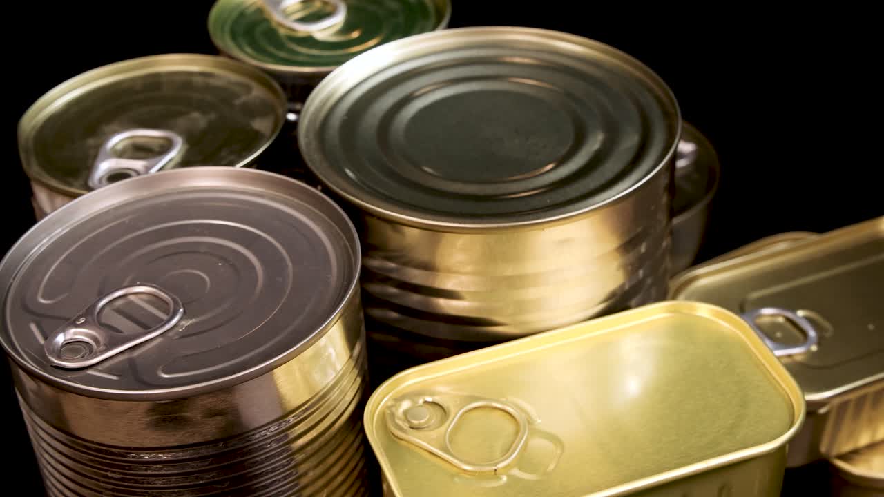 Various unopened canned food containers rotate smoothly under studio lighting, highlighting metallic textures and pull-tab lids against a clean, dark backdrop