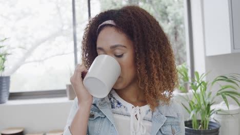 Portrait-of-happy-biracial-woman-enjoying-drinking-coffee-and-smiling-in-kitchen,-slow-motion