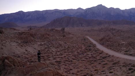 Aerial-drone-view-orbiting-a-man-photographer-standing-on-a-rock-with-a-beautiful-mountain-ridge-and-road-in-the-background-during-blue-early-morning-sunrise-light