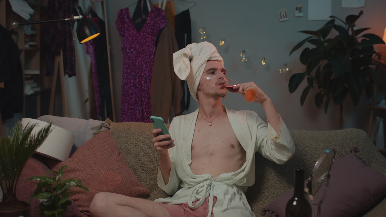 Uomo in accappatoio e asciugamano che si gusta un bicchiere di vino e usa lo smartphone a casa
