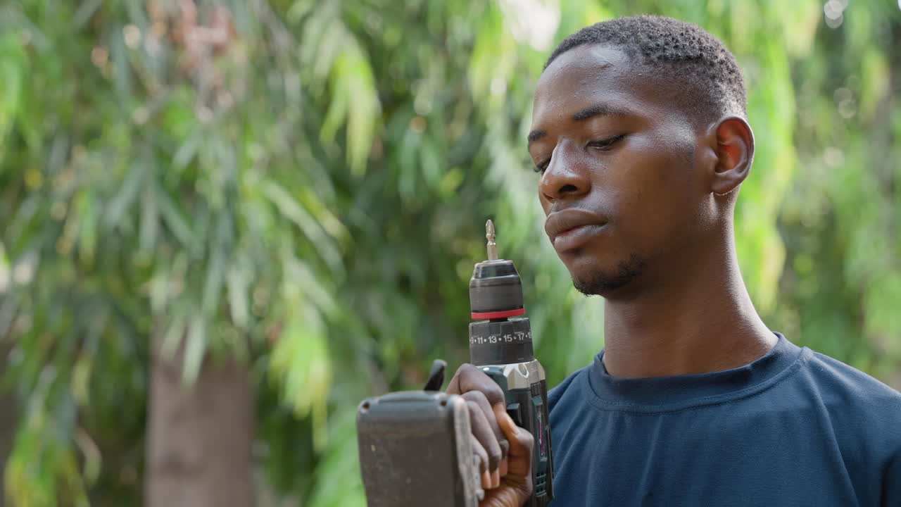 Black carpenter inspecting cordless drill, outdoor leafy background, sweat on face, focused expression checking drill bit and trigger, preparing woodworking project, backyard workshop atmosphere