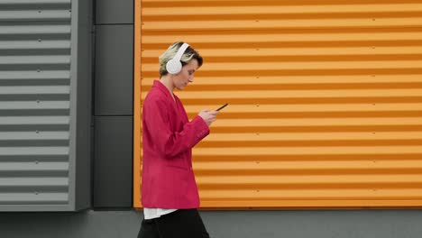 mujer caminando por la calle de la ciudad contra la pared gris y naranja usando un teléfono inteligente.