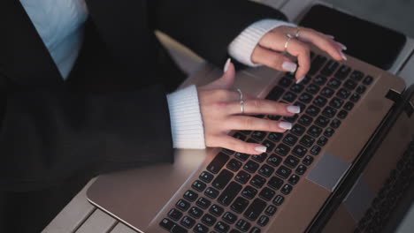 close-up of hands with polished nails typing on laptop keyboard, adorned with silver rings and cozy sweater sleeves, the laptop is placed on a sleek wooden table in an outdoor workspace