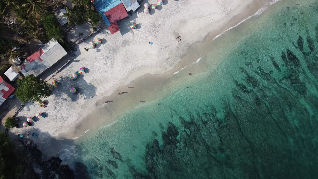 Drone shot showing clear turquoise sea, white sandy coast, beach umbrellas, and small houses in Bali, capturing sunny tropical vibes with people relaxing near gentle waves and vibrant shoreline.