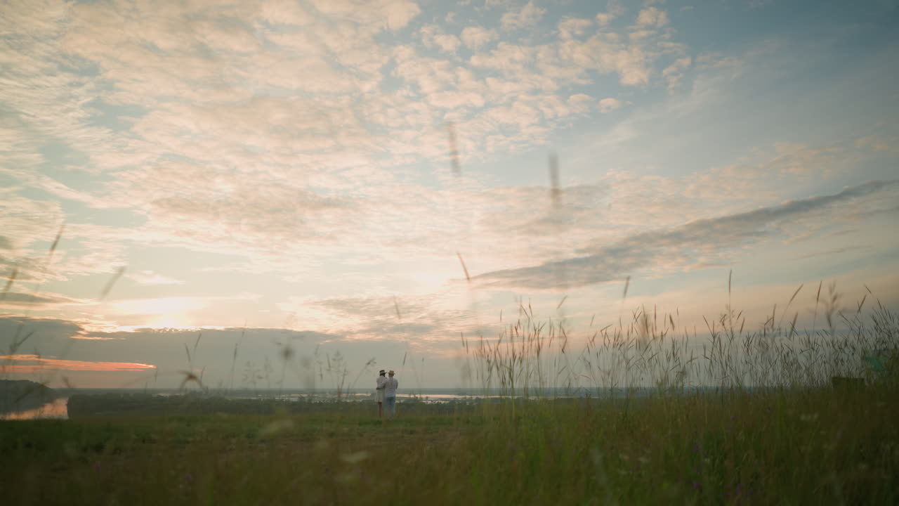 Partners stand on a grassy hill beside a lake at sunset, gazing at the sky. The man, in a white shirt, hat, and jeans, lovingly holds the woman by the waist. She wears a black hat and a white dress