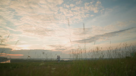 partners stand on a grassy hill beside a lake at sunset, gazing at the sky. the man, in a white shirt, hat, and jeans, lovingly holds the woman by the waist. she wears a black hat and a white dress