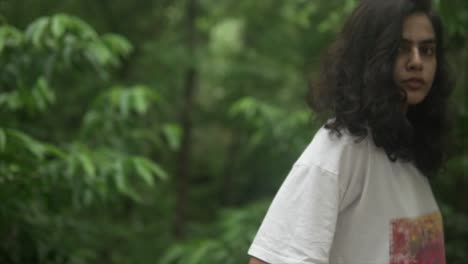 Curly-haired-tan-woman-in-forest-gazes-eerily-tilting-head-in-curiosity