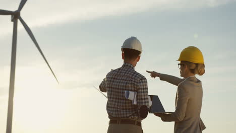 Rückansicht-Von-Kaukasischen-Ingenieurinnen-Und-Ingenieuren,-Die-Einen-Helm-Mit-Laptop-Tragen,-Während-Sie-über-Den-Betrieb-Der-Windmühlen-Turbinen-Sprechen