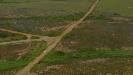 High-angle-view-of-white-car-passing-dirt-road-crossroad.-Tilt-up-reveal-landscape-with-green-vegetation.-South-Africa