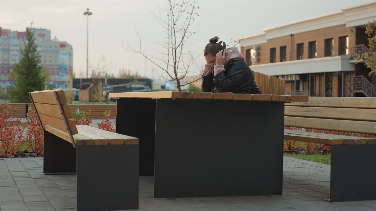 Lady in leather jacket seated at wooden outdoor table putting on headphone surrounded by modern buildings and swaying shrubs on windy day in urban setting
