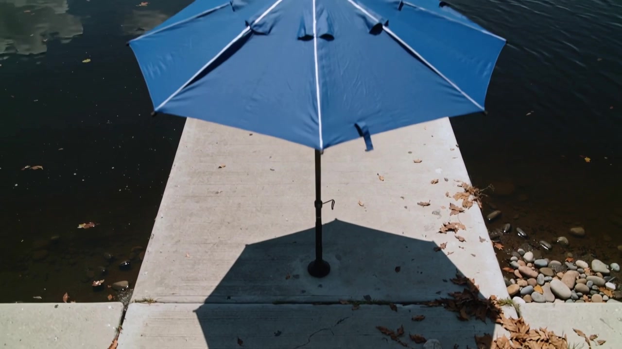 Telecamera che si allontana, rivelando un ombrellone blu da patio posizionato al centro di un molo di cemento, esponendo la costa.