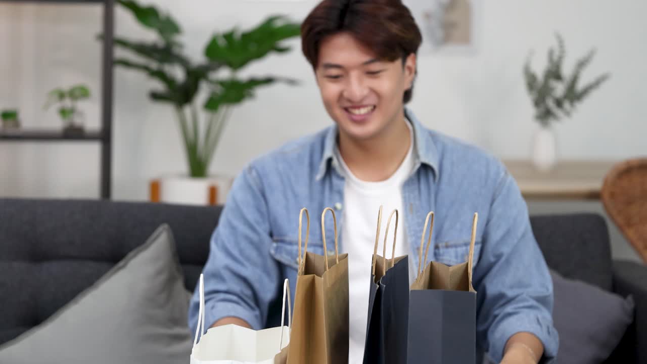 明るい自然光が差し込むリビングルームで、デニムジャケットを着た笑顔の男性がショッピングバッグを開封している。