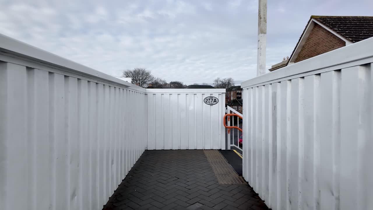 This POV shot shows a footbridge with white metal walls and a gray brick path under an overcast sky