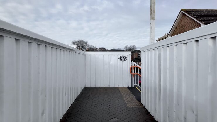 This POV shot shows a footbridge with white metal walls and a gray brick path under an overcast sky