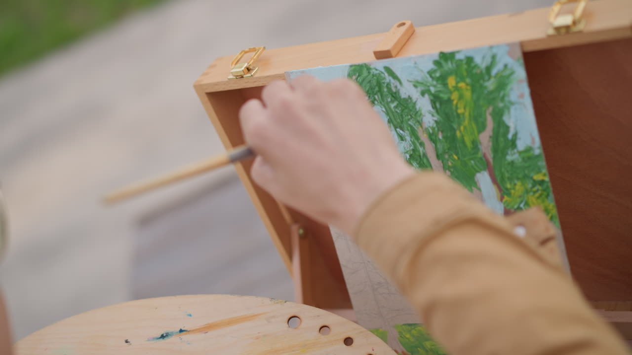 lukisan close-up detail pohon dengan kuas, seorang seniman menyempurnakan sapuan dedaunan pada kanvas kecil di dalam kotak penyangga portabel, palet yang ditutupi dengan warna hijau dan biru, stippling dan blending cepat, fokus di luar ruangan