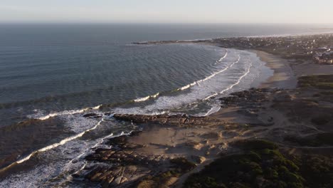 Bella-Toma-Aérea-Que-Muestra-La-Playa-Del-Rivero-Durante-La-Puesta-De-Sol-En-Punta-Del-Diablo,-Uruguay---Olas-Del-Océano-Que-Llegan-A-La-Playa-De-Arena-Y-La-Costa