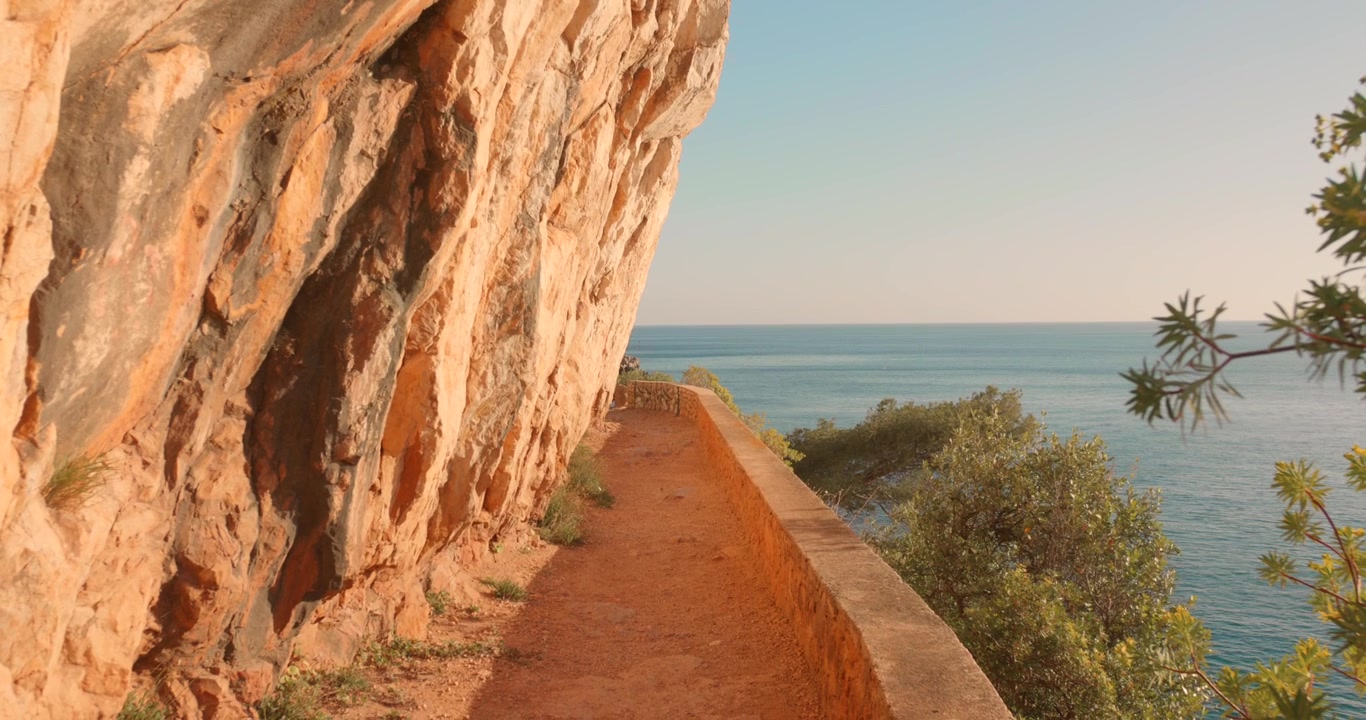 Trilha de terra estreita margeando falésias ocres dramáticas acima do Mar Mediterrâneo. Vegetação costeira exuberante e água turquesa visíveis.