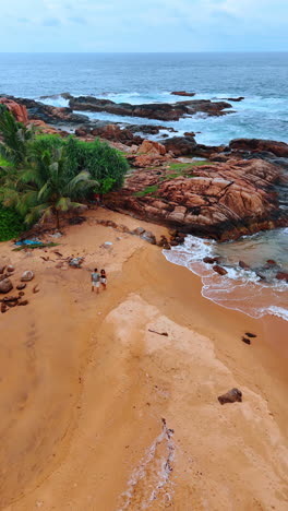 a couple walking by the picturesque sandy beach ending with rocks. ocean waves splash by the boulders. vertical video.
