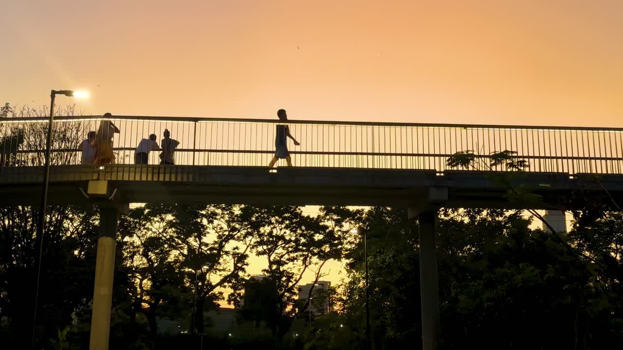 Silhouettes of people walking on a bridge against a vibrant orange sunset sky, surrounded by tree outlines.