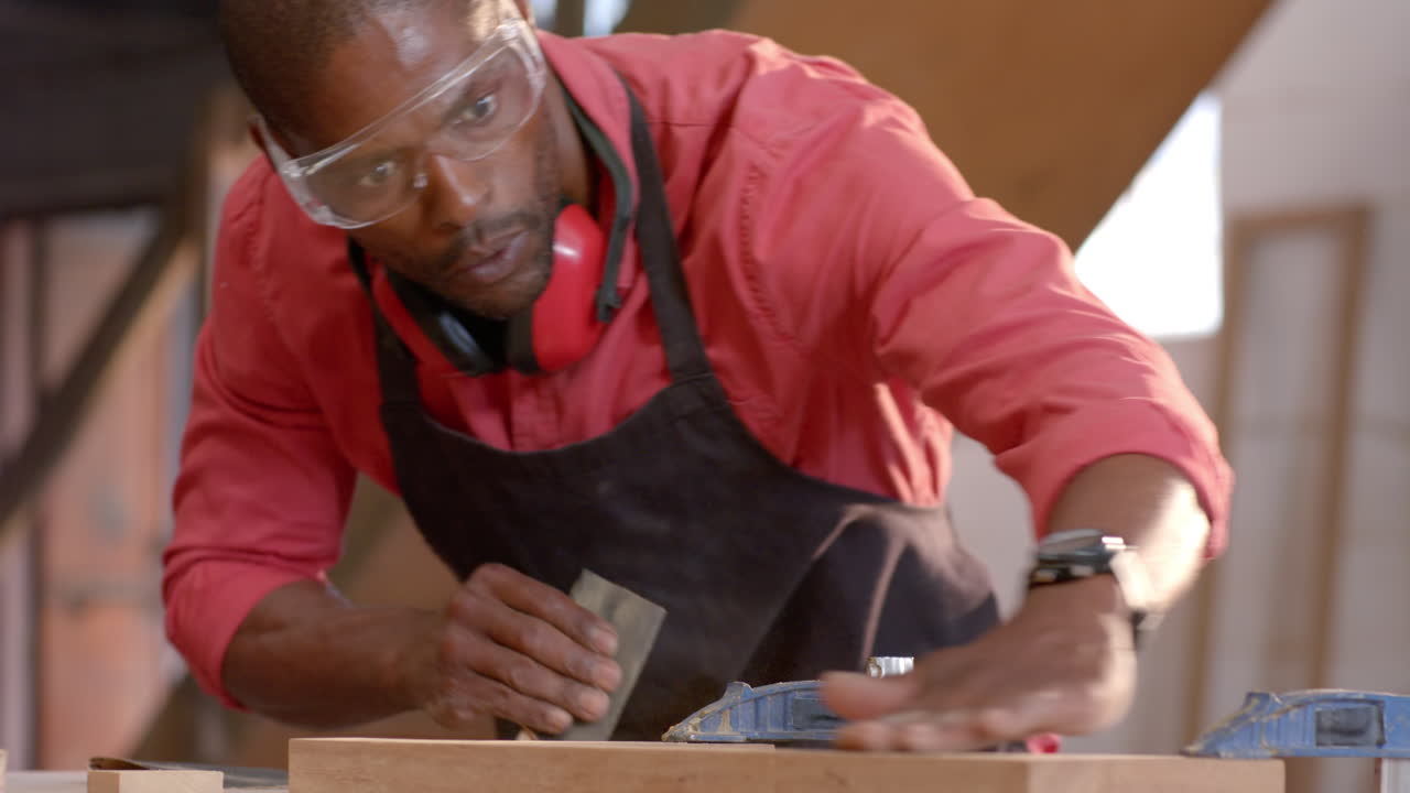 Sanding African American man pressing sandpaper against plank at workbench, with safety goggles
