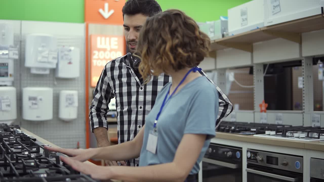 Dark haired, smiling man in plaid shirt with headphones choosing a new modern oven in store. He is planning to buy furniture and home appliances, checking and advising with female consultant