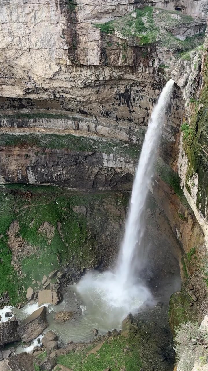 Una potente cascata Tobot scende a cascata da un'imponente parete rocciosa in un bacino roccioso sottostante, circondata da una vegetazione verde lussureggiante