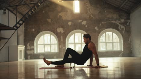 a man in a black sports summer uniform stretches his leg muscles with a special fitness cone in a sports brick hall, which is lit by the rays of the sun