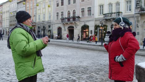 senior wife posing for camera laughing while husband is taking photo pictures on winter city street