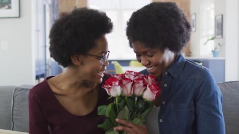 Happy-african-american-mother-and-daughter-sitting-on-sofa,-giving-roses